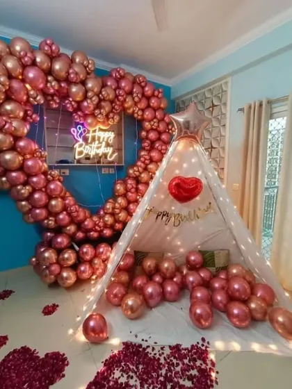 A cozy and romantic birthday surprise featuring a large heart-shaped balloon arch in metallic pink. A small canopy tent and scattered rose petals complete this intimate home setup.