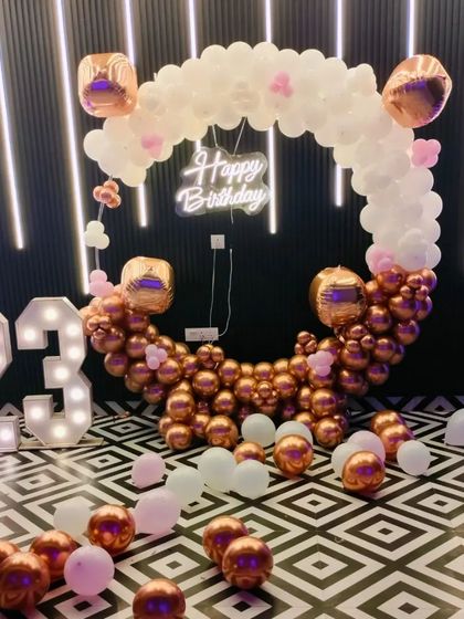 A stylish 23rd birthday celebration with a rose gold and white color scheme. The setup includes a circular balloon arch, a neon sign, and large light-up marquee numbers.