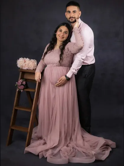 A classic couple's maternity portrait. The partner stands behind the mother-to-be, both gently holding the bump, symbolizing their shared journey and support for one another. The soft pink tones add a romantic feel.