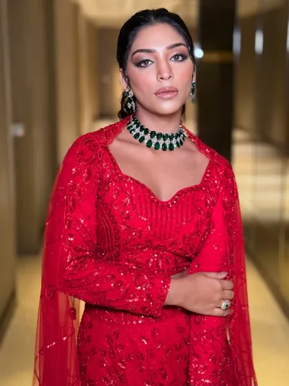 A full-length view of a model in a striking red lehenga. The emerald green choker provides a beautiful color contrast, making this a standout look for any festive evening.