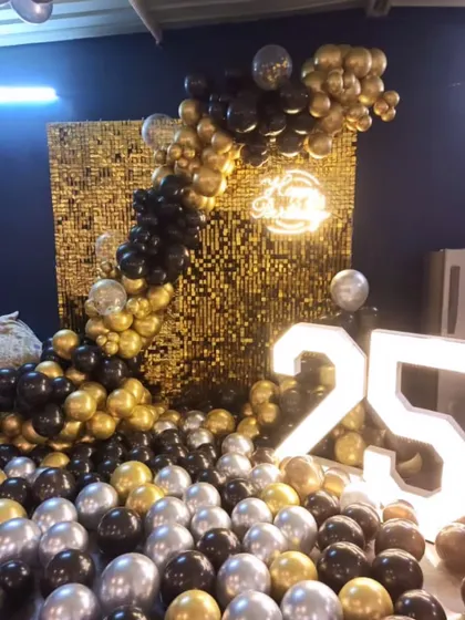 A glamorous 25th birthday or anniversary setup with a golden sequin wall and a cascading arch of black, gold, and silver balloons.