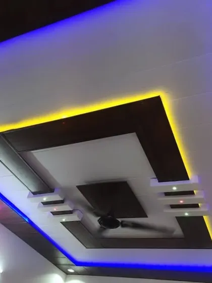 This multi-layered PVC ceiling uses a combination of dark wood and white panels. The dual-color blue and yellow lighting highlights the geometric shapes for a dynamic look.