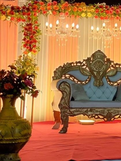 A classic wedding reception stage with a red carpet. The backdrop of peach and white drapes is accented with floral arrangements and chandeliers for a warm and elegant look.