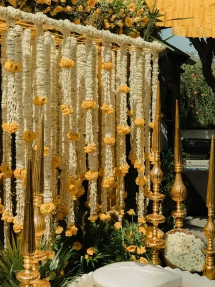 A close-up of an elegant Haldi decor detail. This setup features a wall of white tuberoses and marigold flowers, accented with tall, traditional brass lamps.