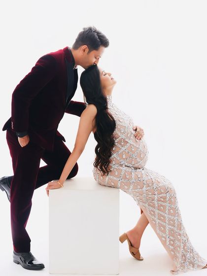A creative and artistic pose that highlights the beautiful silhouette of the baby bump. The contrast between the white background and the formal wear is stunning.