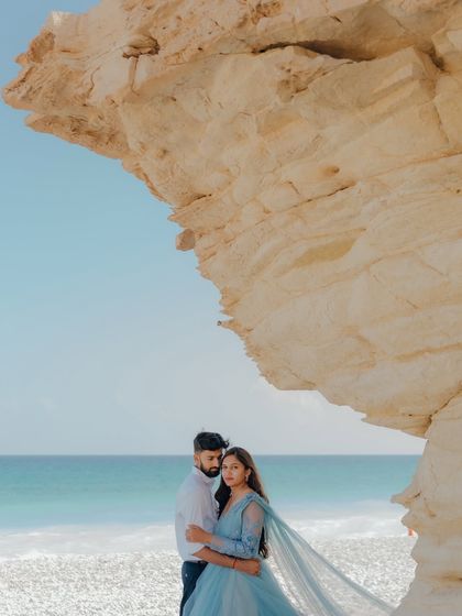 A breathtaking view on the coast of Muscat, Oman. The couple stands beneath a natural rock arch, the turquoise water and blue sky creating a stunning, vibrant backdrop for this serene pre-wedding portrait.
