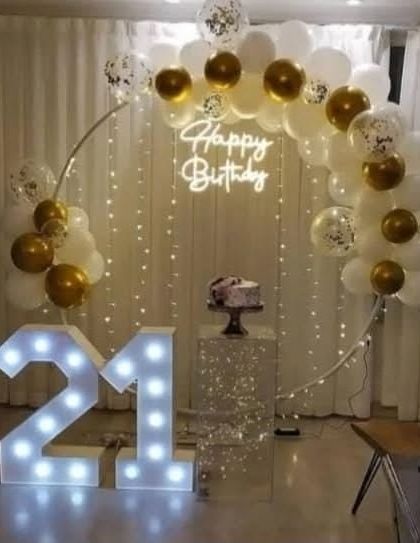 A beautiful 21st birthday setup with a white and gold balloon arch. The decor is completed with a clear pedestal for the cake, a neon sign, and light-up numbers.
