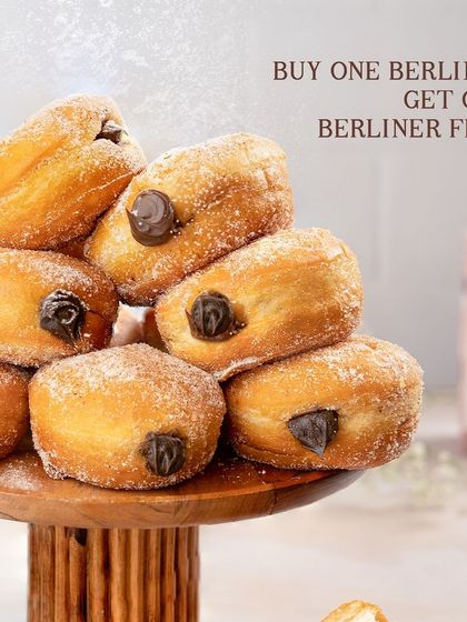 Double the Berliners, double the fun. A beautiful shot of our Nutella and chocolate-filled Berliners stacked high on a wooden stand, showcasing a special BOGO deal.