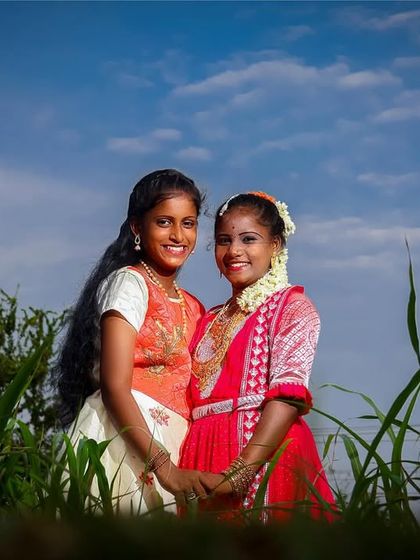 A lovely photo of two young women in traditional attire, sharing a happy moment during an outdoor puberty shoot. The low angle and tall green grass create a beautiful, immersive frame.