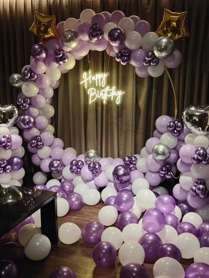 A beautiful purple and white balloon arrangement for a birthday. The setup includes a circular arch, star and heart-shaped foil balloons, and a 'Happy Birthday' neon sign.