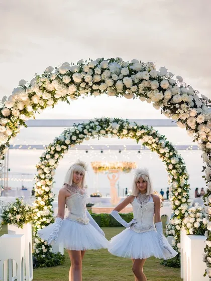 Performers dressed in white, angelic costumes stand at the entrance of a floral archway, welcoming guests to an elegant evening celebration. Their outfits perfectly complement the all-white decor theme.