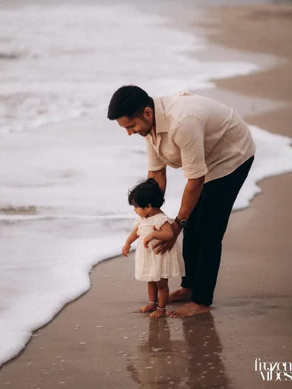 A father and his young child stand at the edge of the ocean, watching the foam of the waves. It's a quiet, gentle moment of introduction and wonder, perfect for a father-child portrait.