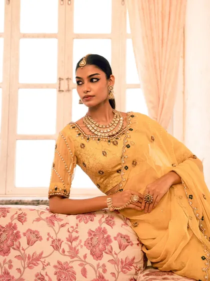 A classic portrait for an ethnic wear brand, focusing on the rich color and detailed craftsmanship of the yellow lehenga and blouse. The floral sofa adds a touch of vintage charm.