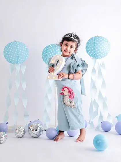 A proud two-year-old holds up her birthday number. The mermaid-themed outfit and pastel blue decorations make this a whimsical and memorable second birthday portrait.