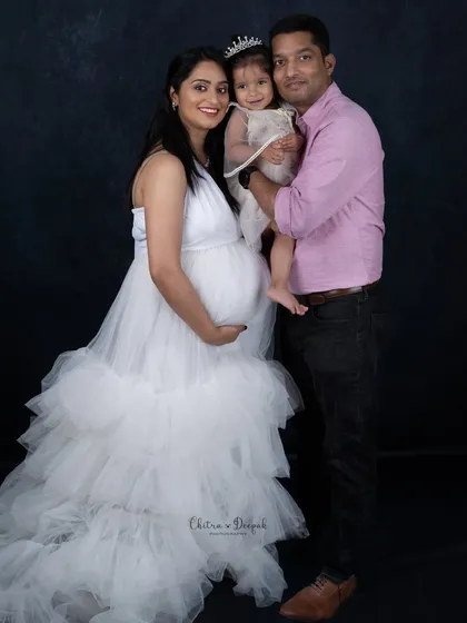 An elegant family portrait with the expecting mother in a flowing white gown, posing with her husband and their little girl.