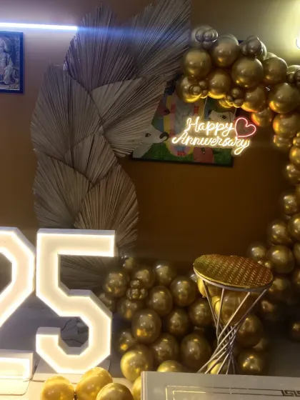 This unique 25th-anniversary setup combines modern and rustic elements. A half-arch of metallic gold balloons is accented with dried palm leaves, framing a "Happy Anniversary" neon sign, with large light-up "25" numbers completing the look.