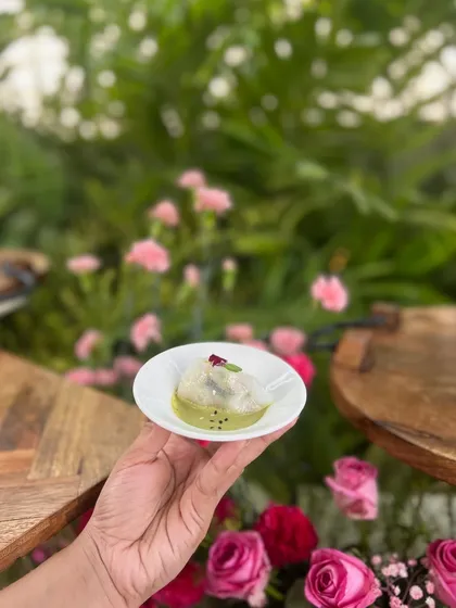 A delicate Thai Basil Dumpling, held in hand against a backdrop of pink roses, from a poolside Haldi celebration.