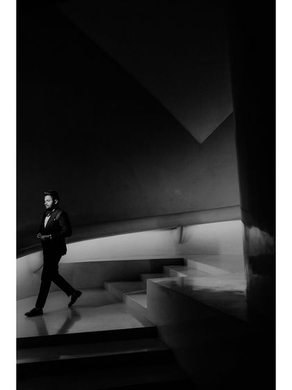 A dramatic black and white shot of the groom, Shrey, walking down a modern staircase. The architectural lines and strong contrast create a powerful and cinematic portrait.