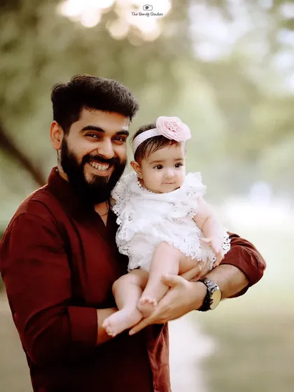 A proud father holds his baby daughter, both smiling for the camera. This father-daughter portrait highlights their special bond against a soft, natural background.