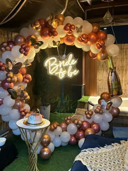 An outdoor terrace setup for a bachelorette party, featuring a circular balloon arch in rose gold and white. The "Bride to be" neon sign glows at the center, creating a perfect photo opportunity next to the jacuzzi.