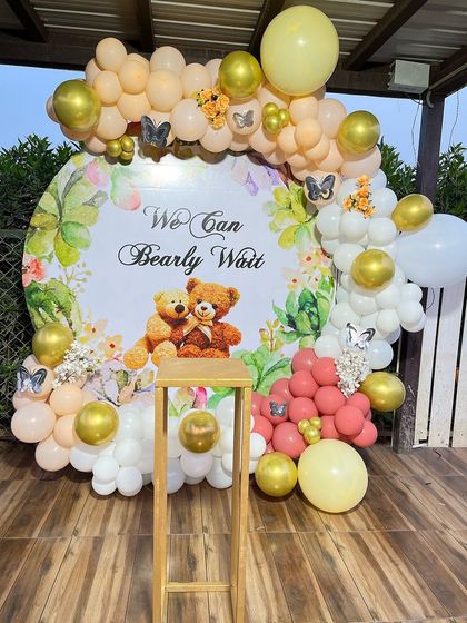 An outdoor "We Can Bearly Wait" baby shower setup. This decoration features a custom backdrop with teddy bears, surrounded by a balloon garland in pastel yellow, peach, and white with butterfly accents.