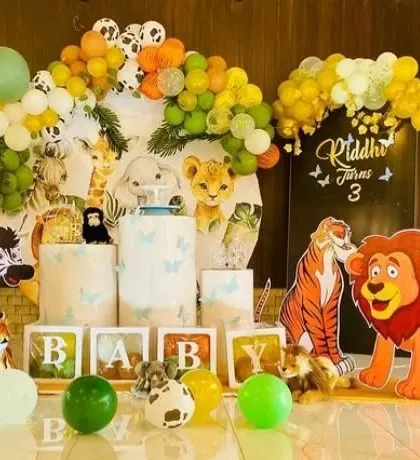 A delightful safari-themed party for Riddhi's 3rd birthday. The setup is filled with cute cartoon animal cutouts like a lion, tiger, zebra, and deer, along with 'BABY' blocks and a green balloon garland.