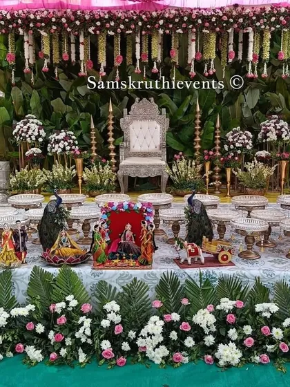 A beautiful baby shower setup with a banana leaf backdrop and hanging floral garlands. The stage is decorated with an ornate silver throne and a full display of offerings on a green lawn.