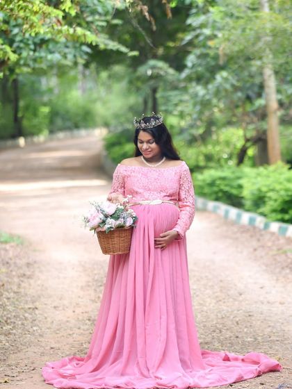 A princess-like portrait of a mom-to-be in a pink gown and tiara, holding a basket of flowers on a country path.