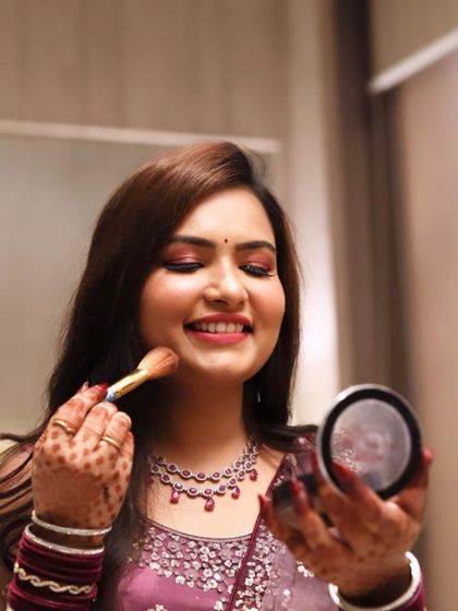 A candid moment during a Roka ceremony makeover. The client is touching up her dewy, fresh makeup, featuring pink eyeshadow and a ruby necklace.