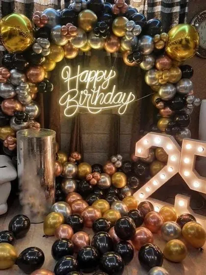 A stylish 25th birthday celebration at home. The decor includes a balloon arch in black, gold, and copper tones, a 'Happy Birthday' neon sign, and a '25' marquee light.