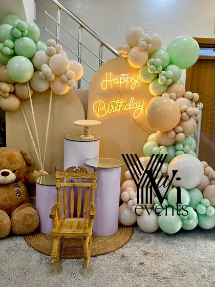 A soft and gentle theme for a first birthday, using pastel green and peach balloons, a teddy bear, and a small throne for the birthday baby.