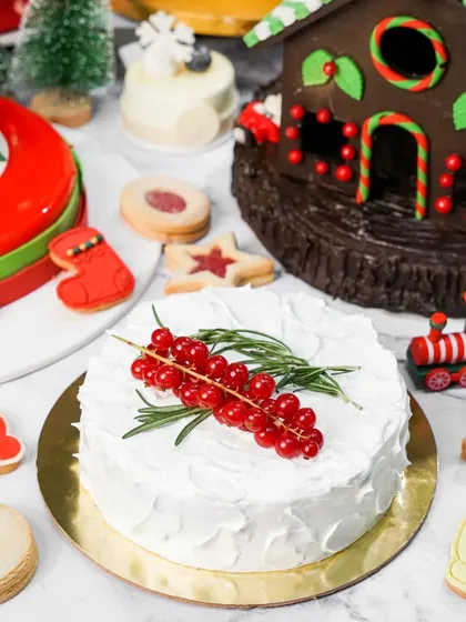 A simple yet elegant white Christmas cake topped with fresh redcurrants and rosemary. The surrounding festive decorations create a beautiful and classic holiday scene.