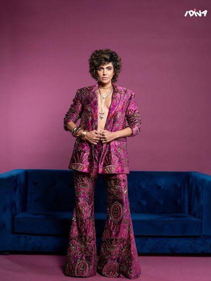 A full-length portrait for iDiva, showcasing the flared pants and bold silhouette of the outfit against a contrasting blue sofa.