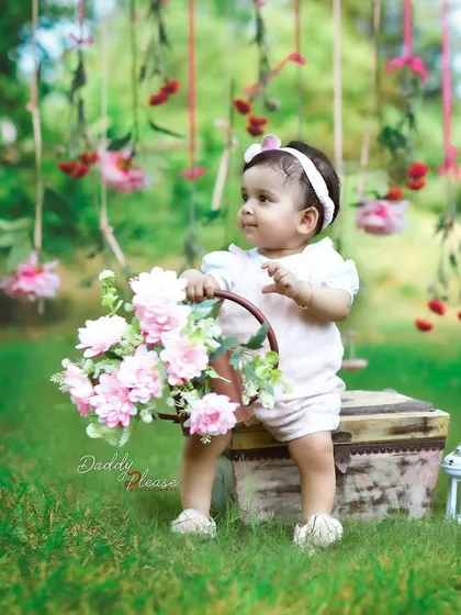 An outdoor sitter session in a beautiful green park. This little girl is sitting on a vintage-style trunk holding a basket of pink flowers, with a lovely floral backdrop hanging from the trees.