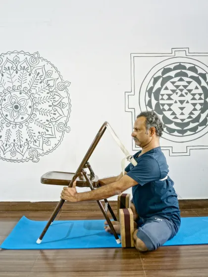 Baddha Konasana (Butterfly Pose) with chair support. This setup helps to open the hips and improve posture while keeping the spine supported.