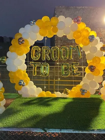 A 'Groom to Be' party backdrop set up on a terrace. This features a circular arch of yellow and white balloons on an artificial grass base, with foil letters spelling out the occasion.