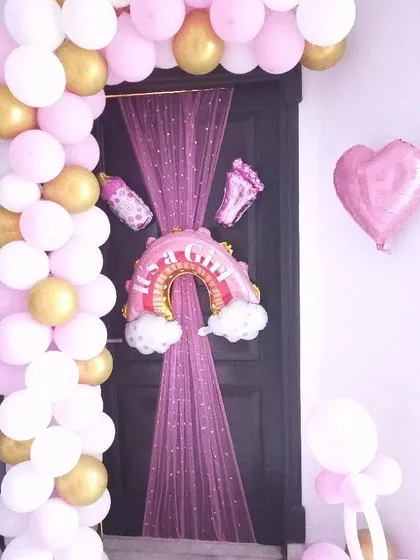 A door decorated to welcome a baby girl, featuring a pink and gold balloon arch and a rainbow foil balloon.