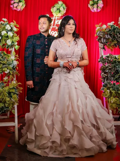 A full-length portrait of the couple at their Roka ceremony. They stand against a festive red backdrop with floral arrangements, a classic shot that captures their coordinated outfits and the celebratory atmosphere.