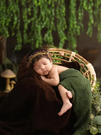 A baby with a delicate leaf crown sleeps in a basket, draped in green and brown blankets, against a backdrop of hanging ivy for a forest-inspired portrait.