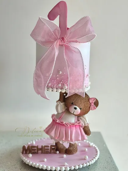 Meet one of our most loved gravity-defying designs. This adorable teddy bear, dressed in a handmade pink frock, holds up a cake for a little one's first birthday. Every detail, from the tiny bow to the delicate pearls, is crafted by hand to make your celebration extra special.
