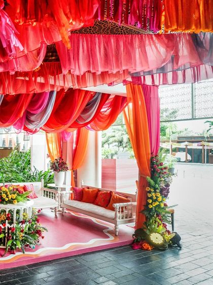A wider view of the Mehendi lounge area, showing the colorful draped canopy, white traditional-style seating with bright orange cushions, and abundant floral arrangements.