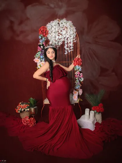 Feeling like royalty in our studio. This mom-to-be wears a crown and a gorgeous red mermaid-style gown, seated in our floral-adorned chair for a truly regal maternity portrait.