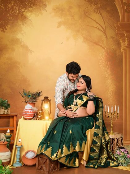 A regal couple's portrait in a traditionally styled set. The mom-to-be looks radiant in a dark green and gold saree, capturing a moment of quiet connection.