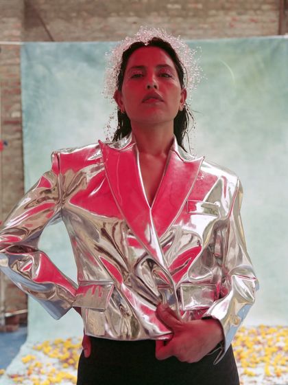 A high-impact editorial look featuring a futuristic, reflective silver jacket. The styling is bold and conceptual, with a delicate, beaded headpiece adding a touch of softness to the sharp, metallic silhouette.