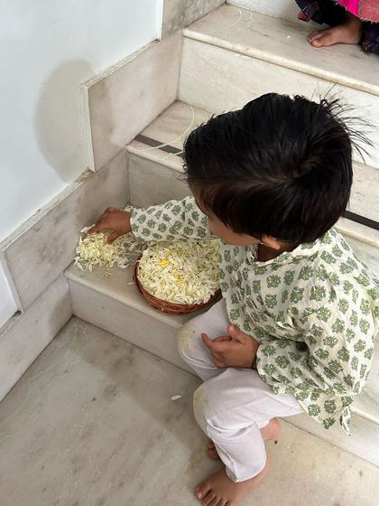 A young child sits on the steps, carefully sorting through a basket of white flower petals, fully engaged in the preparation work.