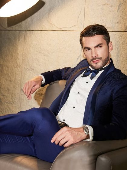 The rich texture of the navy blue tuxedo is evident in this relaxed, seated pose. It demonstrates the comfort and style of a suit made from premium international fabrics.
