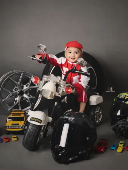 A toddler enjoying his biker-themed photoshoot, sitting on a miniature motorcycle. The set is complete with helmets, wheels, and toy cars for an authentic motor-themed portrait.
