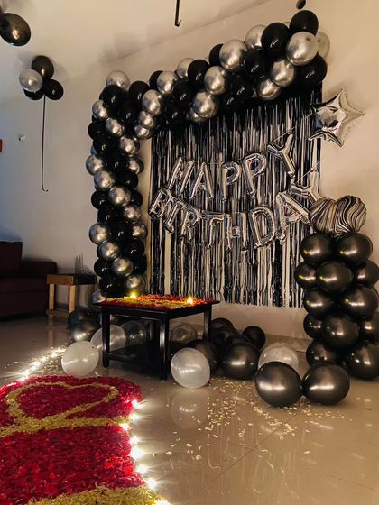 A romantic birthday setup at home with a black and silver balloon arch, a path of rose petals, and candlelight.