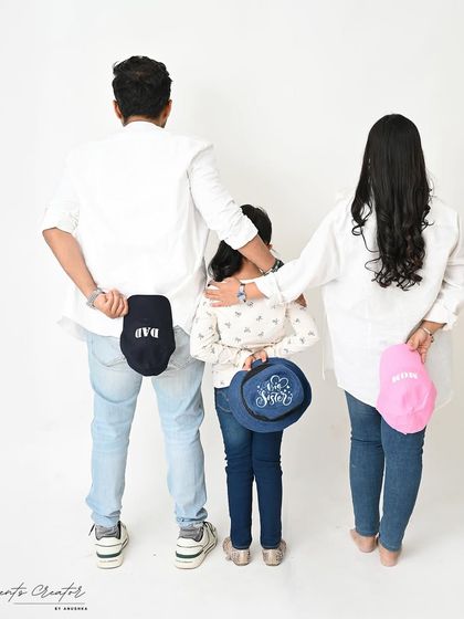 A fun and creative family announcement photo. With their backs to the camera, the family holds hats that reveal their roles: Dad, Mom, Sister, and a tiny pink one for the new baby girl.