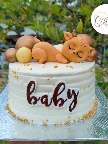 A sweet and simple baby shower cake. This design features an adorable sleeping baby deer figurine nestled among gold and brown baubles on a clean white buttercream cake.